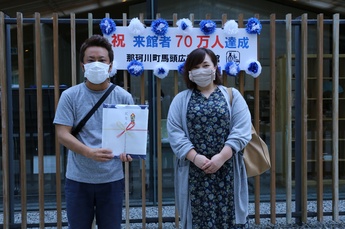 前後賞の1組目、祝来館者70万人達成の看板の前に立つ、左から記念品を持つ男性、女性